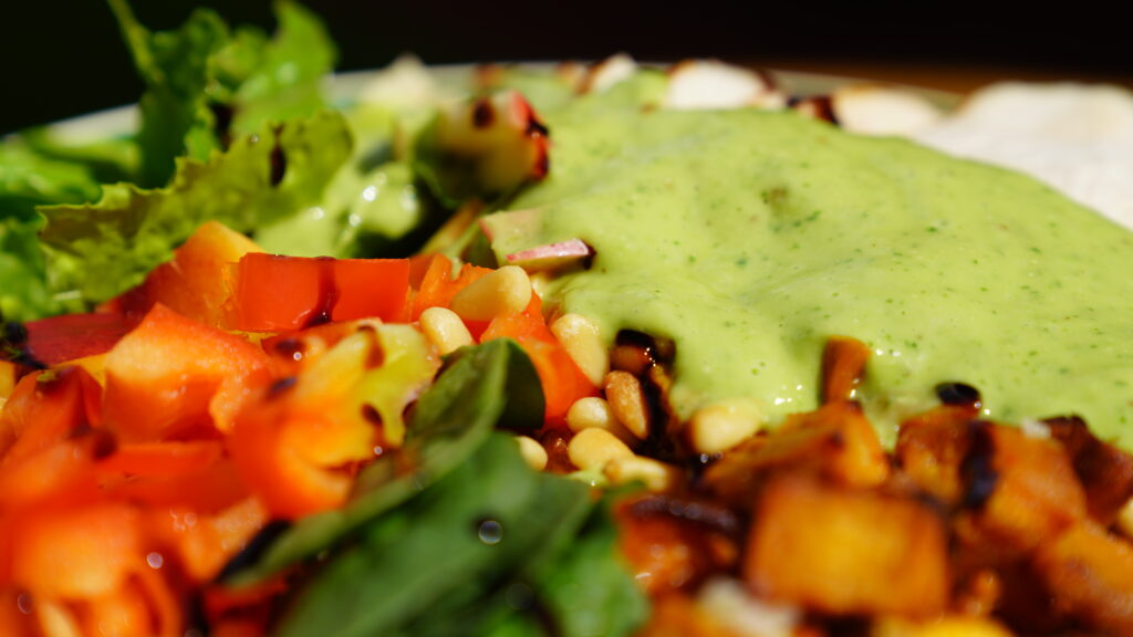 Close-up of a colorful grain bowl with roasted sweet potatoes, red peppers, fresh greens, pine nuts, and creamy green avocado dressing.